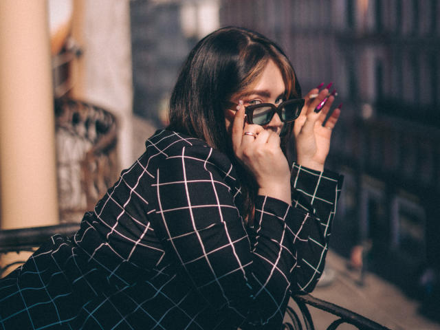 LunaLise with dark brown hair, wearing a black checkered shirt, holding black sunglasses to her eyes, showing off red-painted nails, sitting outdoors on a metal chair.