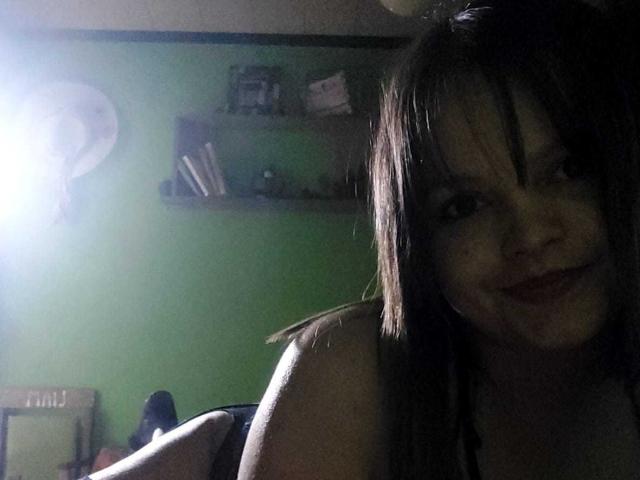 VictoriaKoad with dark brown hair, smiling, wearing a tank top, lying down, visible shoulders and upper chest, soft light, green wall, shelf in background, camera flash on left.