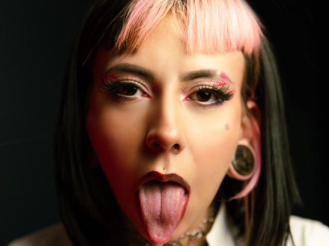 TerriClement with pink and black hair, sticking out her tongue, pink eyeliner, nose piercing, and large gauged earrings, looking directly at the camera with a playful expression.