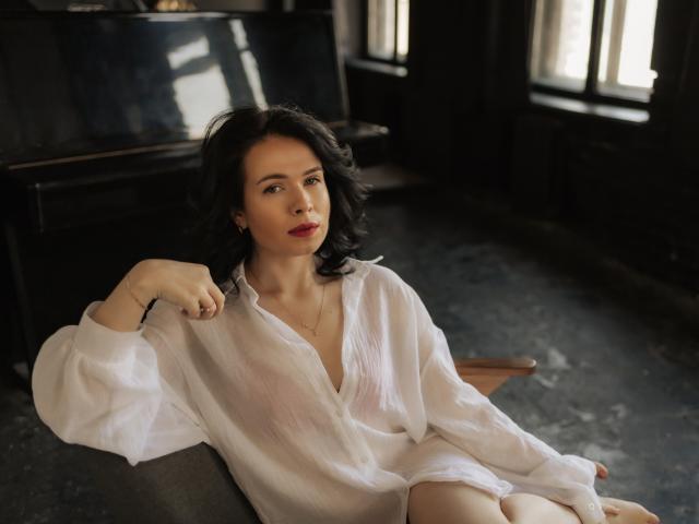 JasmineKouch with curly black hair, wearing a white, slightly sheer button-up shirt, sitting on a gray chair, looking up with red lipstick and a subtle smile.