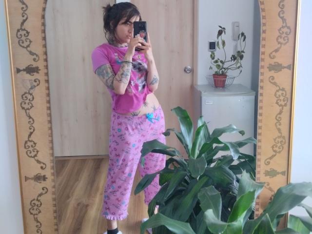 Amy, con tatuaggi, indossa una maglietta rosa a fiori e pantaloni rosa coordinati, e sta scattando un selfie allo specchio. Ha un sedere prosperoso, piccoli seni e, sullo sfondo, una pianta in vaso..