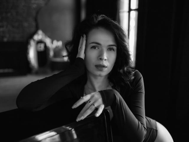 Black-and-white photo of JasmineKouch with wavy dark hair, wearing a long-sleeve top, leaning on a table, hand on her head, looking serious, ring on her finger.