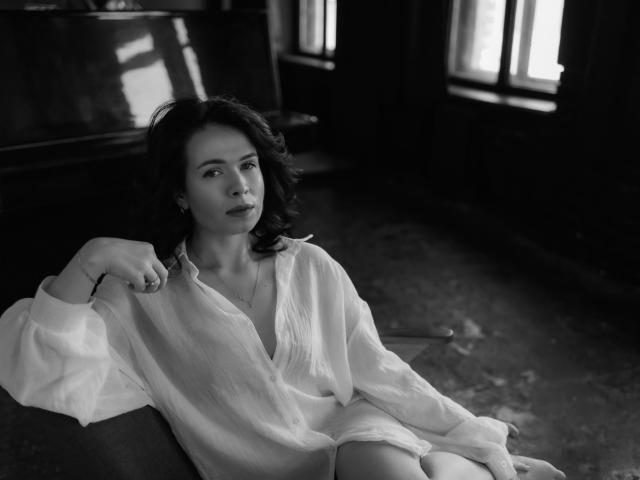 Black-and-white photo of JasmineKouch with medium tits, dark wavy hair, wearing an oversized white shirt, sitting in a dimly lit room, looking to the side with a relaxed expression.