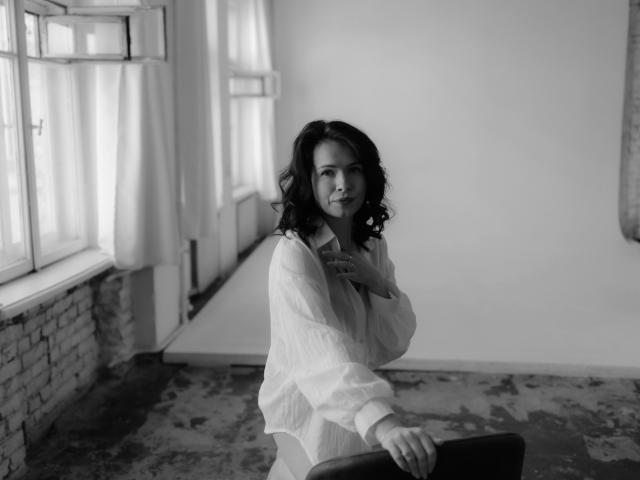 Fotografía en blanco y negro de JasmineKouch con cabello rizado, vestida con una camisa blanca holgada, sentada, con la mano cerca de la barbilla, mirando a la cámara, una pared de ladrillos y una ventana a la izquierda, suelo con diseño..
