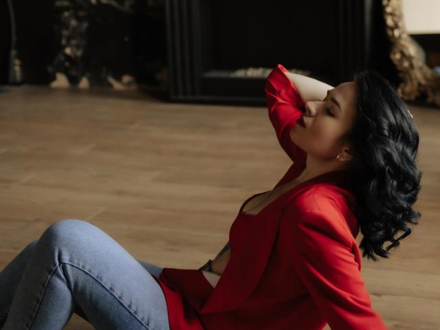 JasmineKouch sitting on wooden floor, back against black fireplace, wearing red jacket, blue jeans, hand touching curly black hair, eyes closed, looking relaxed, red jacket open slightly.