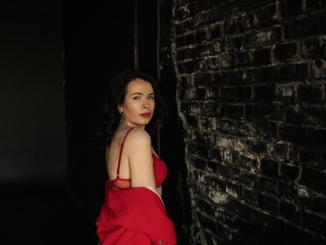 JasmineKouch, com cabelo cacheado escuro, vestido vermelho e alças de sutiã vermelhas, está em pé contra uma parede de tijolos escuros, olhando para trás com um leve sorriso, mostrando seu traseiro..