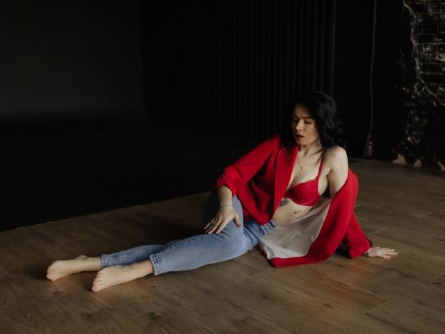 JasmineKouch, with red bra and bare tits, sits on wooden floor, wearing open red shirt and blue jeans, black wavy hair, bare feet, dark background, looking to the side.