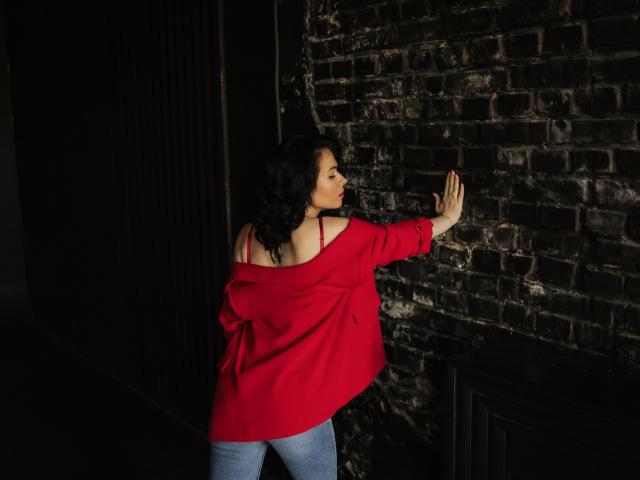 JasmineKouch, con cabello negro rizado, una blusa roja con un hombro descubierto y jeans azules, tocando una pared de ladrillo oscuro con su mano derecha, mostrando su espalda y su figura delgada..