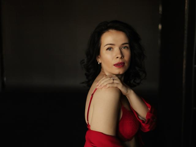JasmineKouch with dark curly hair, red bra, and matching red top, hands on shoulder, slight smile, red lipstick, dark background, looking at camera, sexy, soft lighting.