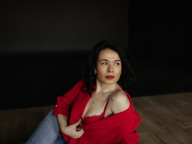 JasmineKouch, avec ses cheveux foncés, son soutien-gorge rouge et sa chemise rouge, dévoilant ses seins, assise sur un plancher en bois, regardant sur le côté, fond noir, plancher en bois, chemise légèrement ouverte..