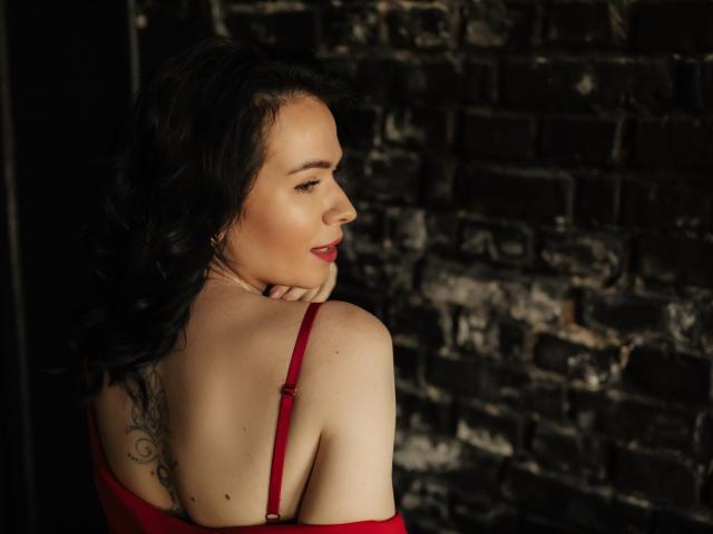 JasmineKouch with wavy black hair, red dress, and tattoo on her back, looking over her shoulder, showing her bare shoulder and red bra strap, smiling with red lipstick.