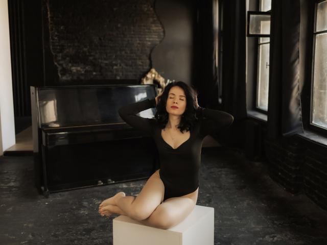 JasmineKouch with pale skin, black curly hair, and red lipstick, wearing a black long-sleeve bodysuit, sitting on a white cube, hands behind head, eyes closed, and legs folded.