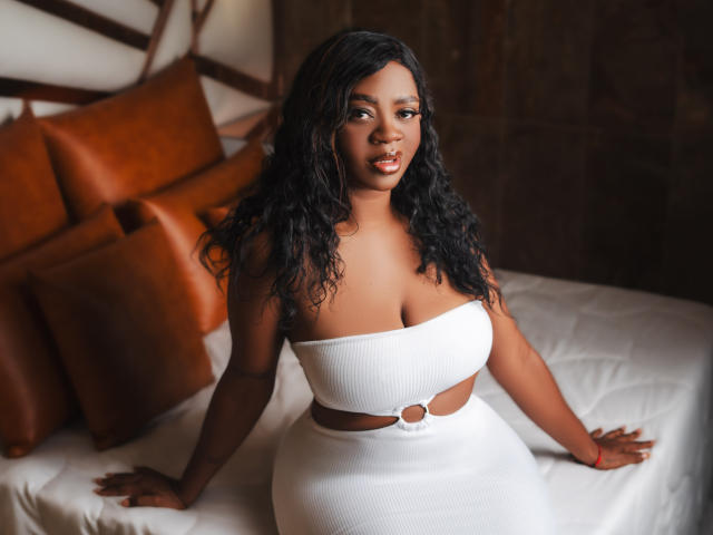 LuisaCastelX with curly black hair, brown skin, and large tits, wearing a white strapless dress with a ring cutout, sitting on a bed with brown pillows.
