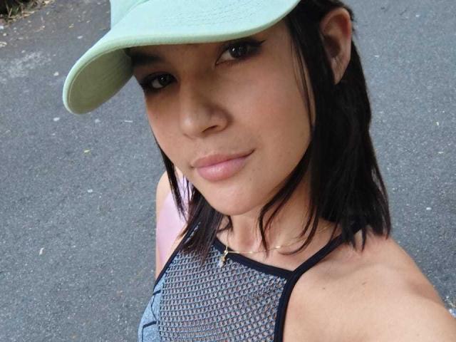 ScarlletZhen with medium tits, wearing a green cap, black mesh top, and a gold necklace, looking up with a slight smile. She has dark hair and light brown skin.