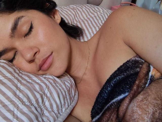 ScarlletZhen sta dormendo con gli occhi chiusi, le labbra rosa e delicate, e una collana d'oro. È coperta da una coperta morbida marrone e nera, con un cuscino a righe bianche..
