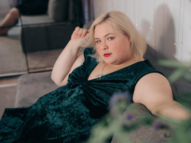 EllaBigBabe, avec sa peau pâle, ses cheveux blonds et son rouge à lèvres rouge, portant une robe en velours vert foncé, allongée sur un canapé gris, tenant ses cheveux et regardant l'appareil photo..