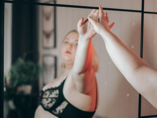 EllaBigBabe's handen die een spiegel aanraken, waarin haar zwarte kanten bh en bleke huid worden weergegeven, met een onscherpe achtergrond. Haar blonde haar is licht zichtbaar..