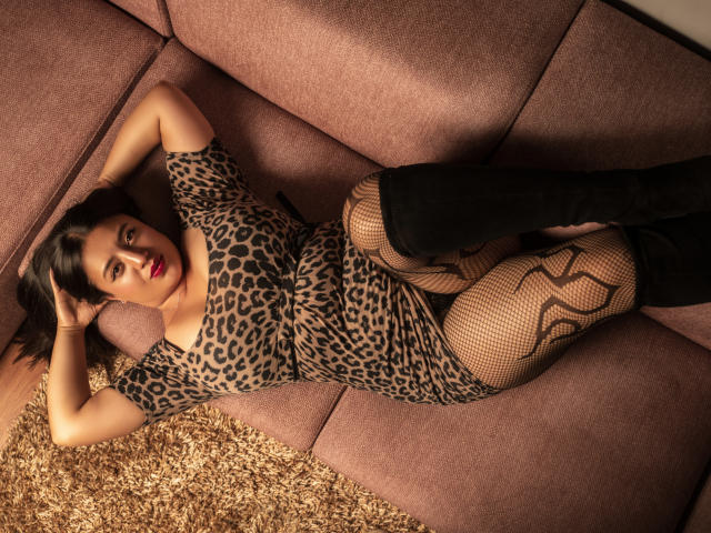 VictoriaVolkov is lying on a pink couch, wearing a leopard-print dress, fishnet stockings, and black knee boots, with her arms above her head and red lipstick.