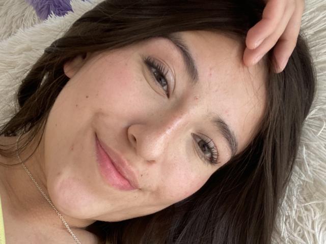 KatiaRogers has light skin, brown eyes, and long brown hair, smiling with a slight blush, touching her head with one hand, wearing a silver necklace.