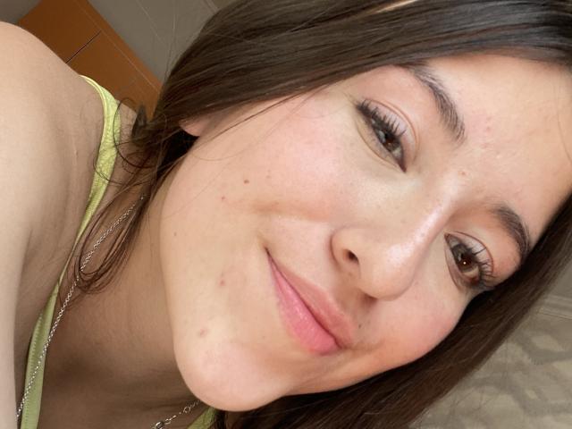 KatiaRogers' face, showing light skin with freckles, brown eyes, long brown hair, subtle smile, small nose, slight blush, wearing yellow top, silver necklace, and small zit on cheek.
