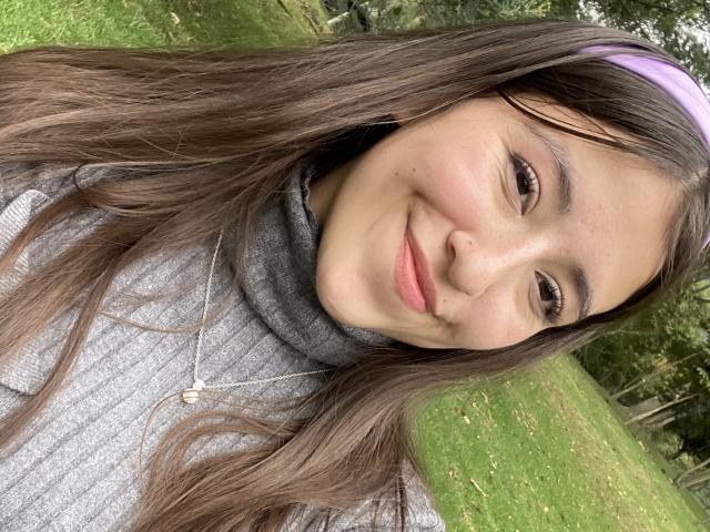 KatiaRogers has brown hair, a purple headband, and a gray turtleneck, smiling softly with a small nose piercing, wearing a silver necklace with a small pendant.