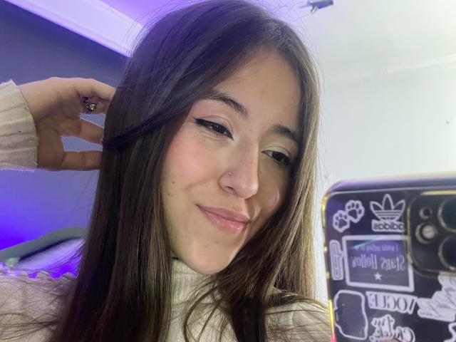 KatiaRogers has long brown hair, a nose piercing, and is smiling at a mirror selfie with her phone, wearing a beige sweater, showing her cute face and small ass.