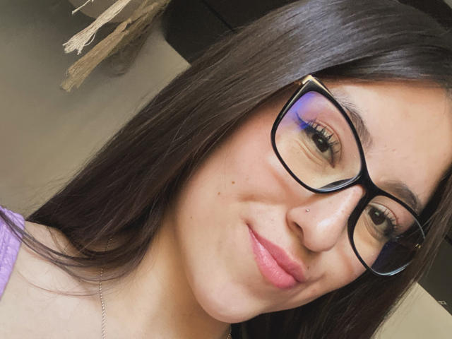 KatiaRogers has long brown hair, a nose ring, and a slight smile. She's taking a selfie with a black phone, wearing a beige sweater, looking cute.