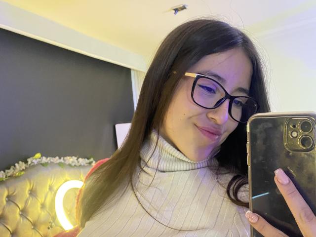 KatiaRogers has dark brown hair, black glasses, pink lips, a nose ring, and a few small moles on her face, smiling slightly. She's wearing a purple top.