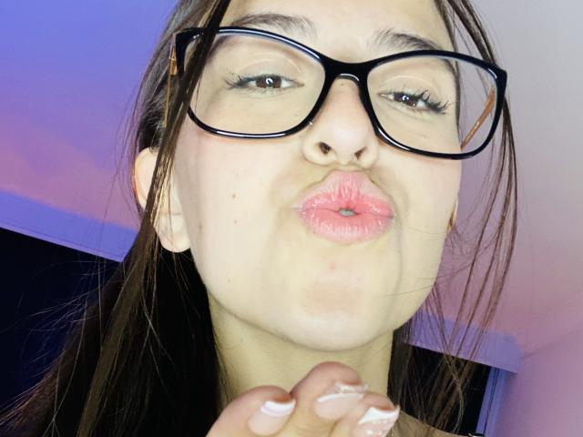 KatiaRogers with long brown hair, glasses, and a white turtleneck, taking a selfie with a black phone in a room with a tufted, yellow headboard.