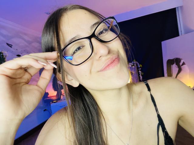 KatiaRogers with pink lipstick, puffy lips, black glasses, brown hair, and white nail polish, making a kissing face, holding her fingers near her mouth.