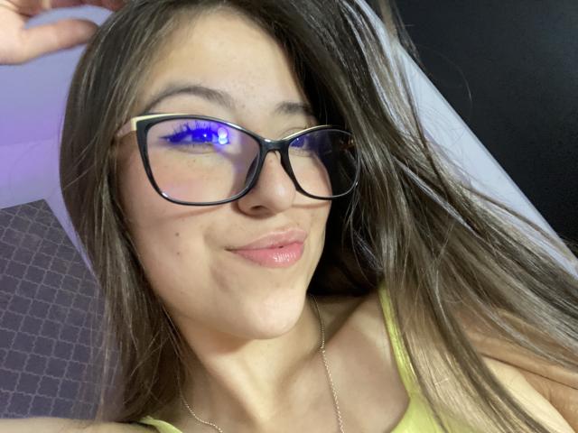 KatiaRogers with long brown hair, glasses, and black strappy lingerie, holding phone, covering small tits with hands, standing in room with Eiffel Tower wall art.