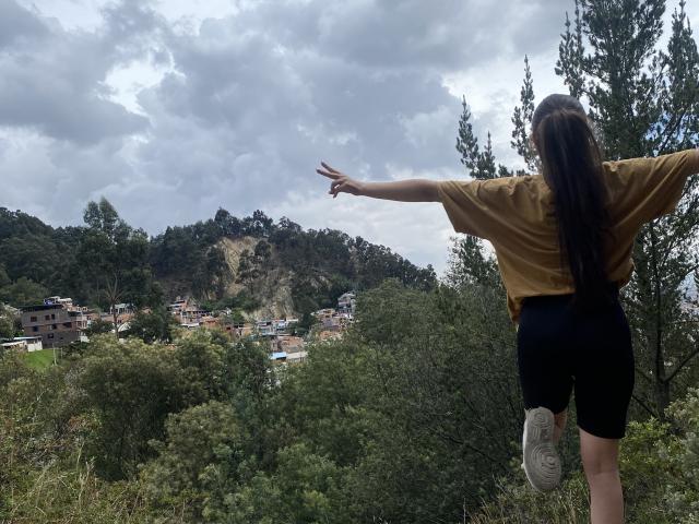 KatiaRogers with a brown shirt and black shorts, standing with her ass facing the camera, looking at a hillside village. She's wearing rainbow socks and has a ponytail.
