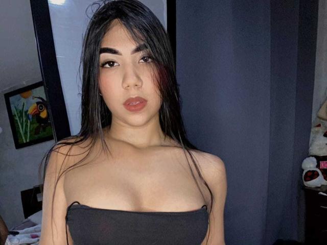 Ariana Murphy, curvilínea e com tatuagens, vestindo uma cropped preta e uma saia preta, está sentada em uma cama, com as pernas cruzadas, mostrando suas nádegas e tatuagens, usando óculos e com um sorriso sedutor..