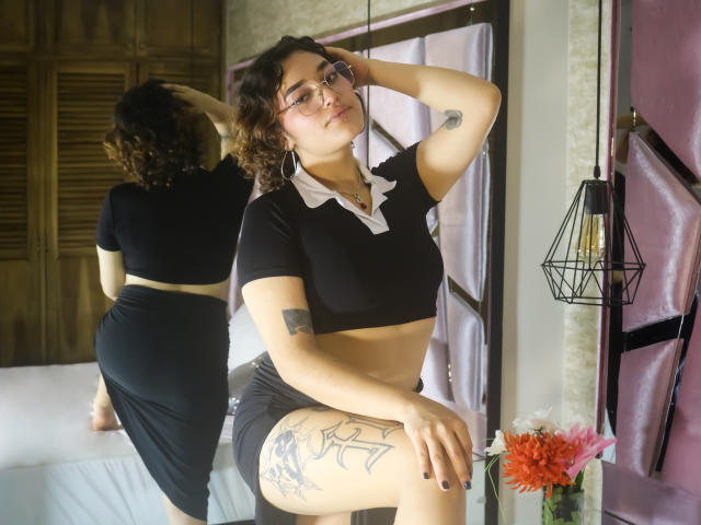 ArianaMurphy with curly brown hair, glasses, and a nose ring, wearing a black crop top, finger on her lip, black skirt, and visible tattoos, sitting on a white couch.