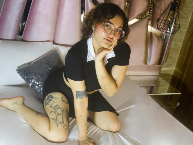 ArianaMurphy with curly brown hair, glasses, black crop top, black skirt, showing tattoos, sitting on bed, hand in hair, reflection in mirror, flowers on right.