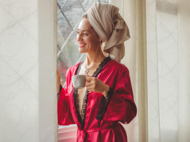 MiaMott with a towel on her head, smiling, holding a white cup, wearing a red satin top with black lace, diamond-patterned window in front.