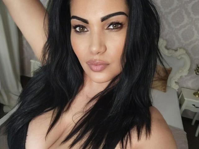BeliseChappelleX-hot has long black hair, brown eyes, thick eyebrows, and is wearing no clothes, showing her tits and pouty ass-pleating lips. Her arm is raised above her head.