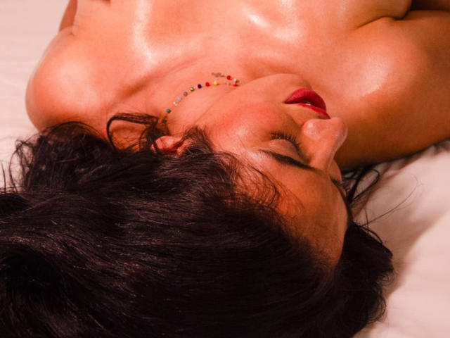 HeraDiosa's shiny, oily tits and red lipstick are visible as she lies face-up with dark hair, eyes closed, and a colorful bead necklace, in a sensual, relaxed pose.