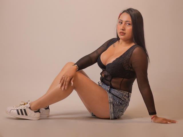MadyBrown met grote borsten, zittend, draagt een zwarte doorschijnende top, denim shorts, witte Adidas sneakers, lang zwart haar, bruine huid, gouden horloge, kijkt naar de camera, beige achtergrond..
