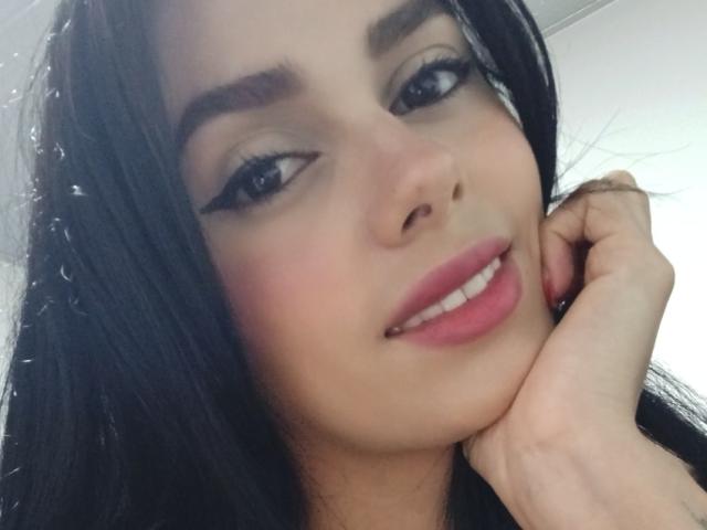 AnaisHilton with black hair, blue eyes, and pink lipstick, smiling, holding her chin with a hand, wearing bold eyeliner, and looking slightly to the side.