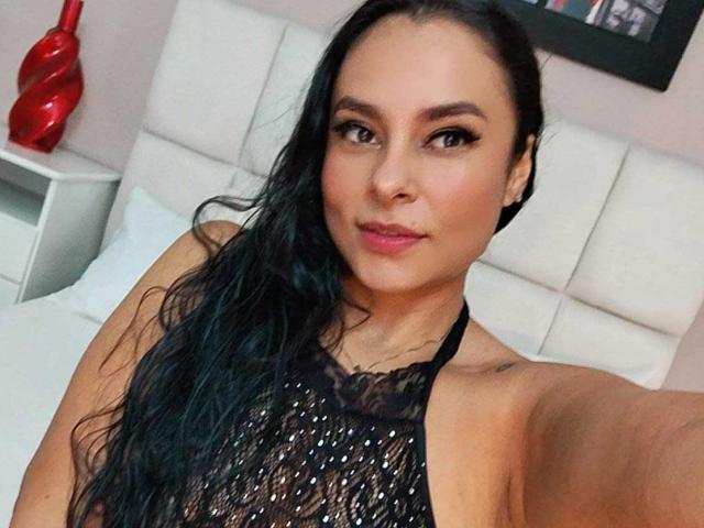LunaSoler with long black hair, wearing a sheer black halter top, showing her tits, and pink lipstick, taking a selfie in a bedroom.