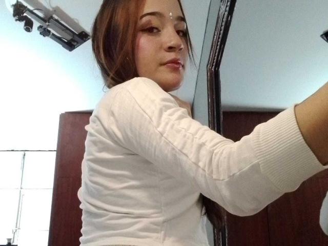 MadisonAngel met bruin haar, een wit shirt met lange mouwen en subtiele make-up, die een selfie maakt, haar gezicht iets gedraaid, haar rechterarm omhoog, de camera is boven haar hoofd bevestigd..