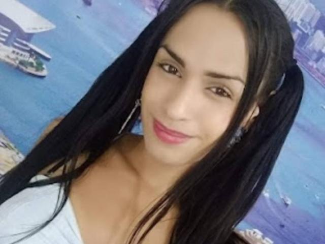 AngieTsSex with black pigtails, brown eyes, pink lipstick, white strapless top, smiling, showing tits, wearing silver earrings, light skin, and dark hair, in a selfie.