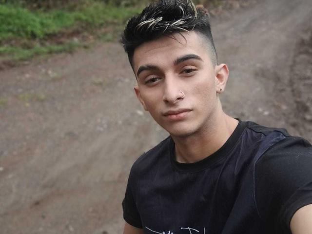 CandyEngel has short, dark hair with blonde highlights, a nose piercing, and a black t-shirt. He's looking down with a slight smirk. He's a cute, sexy young dude.