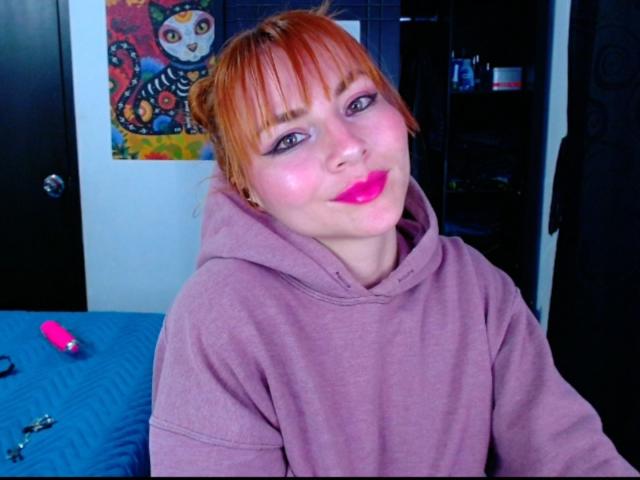 EmelySofia with red-orange hair, pink lipstick, and pink hoodie, sitting, smiling, in a room with a blue bed and colorful cat painting. Vibrant makeup, cute, sexy.