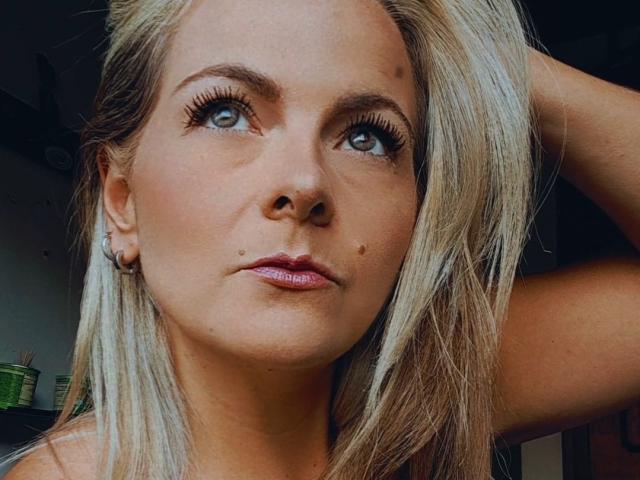 PerlaGoloza has blonde hair, blue eyes, and a small mole on her left cheek. She's looking up, with her right arm raised, showing off her tits.