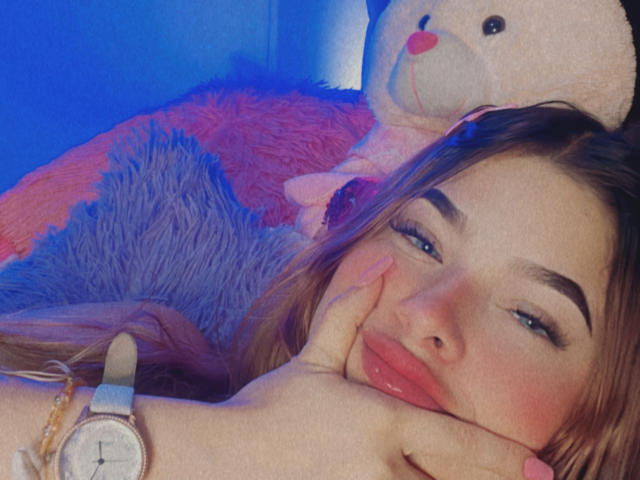 ShantalSummer's face with pouty pink lips, blue eyes, and brown hair, resting on a hand, wearing a watch, with a stuffed teddy bear in the background.