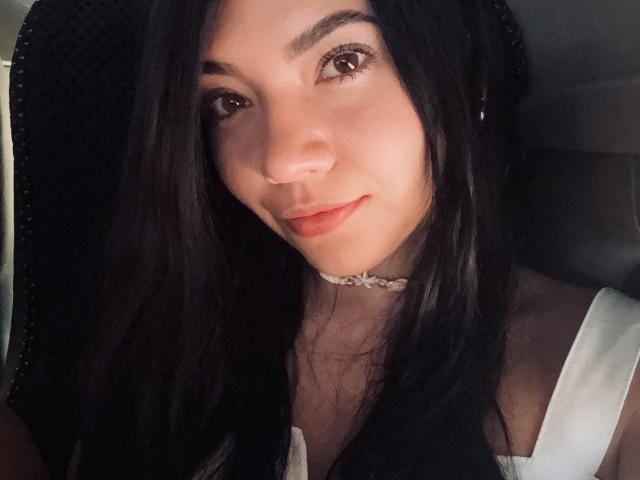 SofieWhite with black hair, brown eyes, and a slight smile, wearing a white top and a braided choker, looking cute and sexy, sitting in a car.