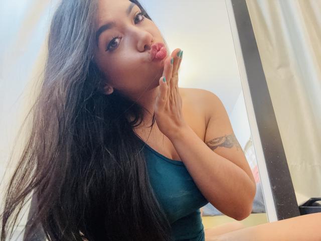 SofieWhite with long black hair, pouting lips, green tank top, tattoo on right arm, and green nail polish, kissing her fingers. Her tits are visible. Mirror reflection.