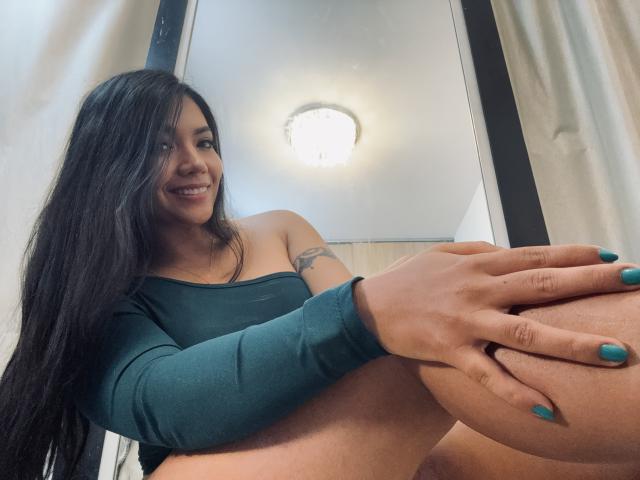 SofieWhite with long black hair, smiling, wearing a green long-sleeve top, holding her big ass with teal nails, visible tattoo on her left shoulder.